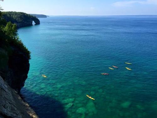 A scenic view of clear blue waters with several kayakers paddling near a rocky shoreline and lush green trees.
