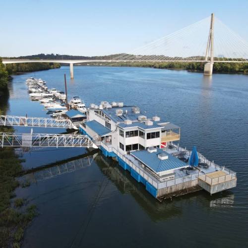 Aerial view of a floating restaurant on a river, with boats docked nearby and a bridge in the background.