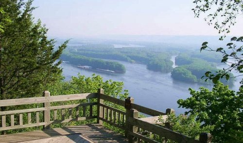 A scenic view from a wooden overlook, showcasing a river winding through lush green trees and hills in the distance.
