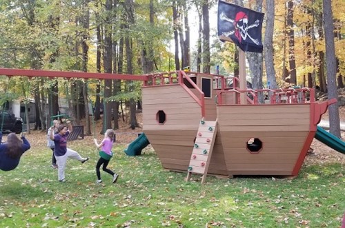 Children play on a pirate ship-themed playground in a wooded area with autumn leaves. Swings and a slide are visible.