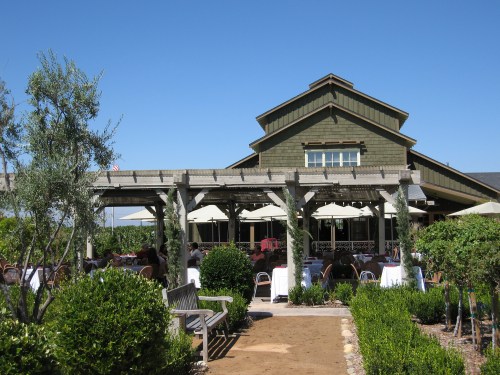 A rustic restaurant with a patio, surrounded by greenery and outdoor seating under umbrellas on a sunny day.