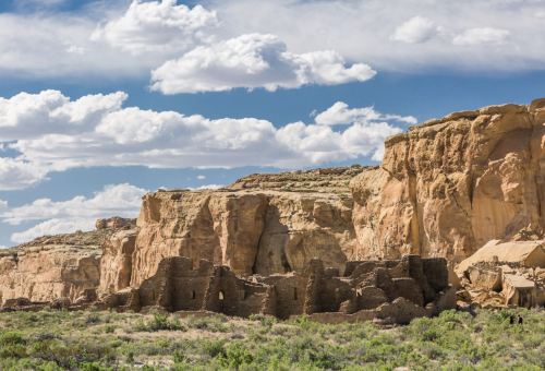 Ancient ruins nestled against a rocky cliff under a blue sky with fluffy clouds, surrounded by green vegetation.