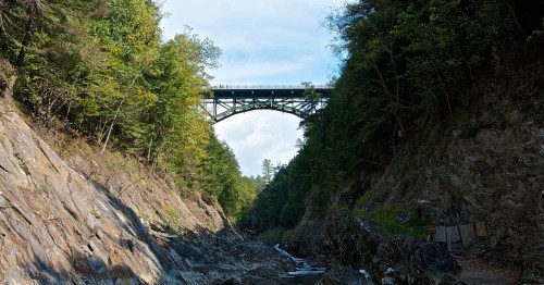 A tall bridge spans a rocky gorge, surrounded by lush green trees under a partly cloudy sky.