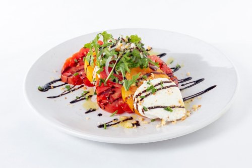 A colorful salad with sliced tomatoes, fresh herbs, and creamy cheese drizzled with balsamic glaze on a white plate.