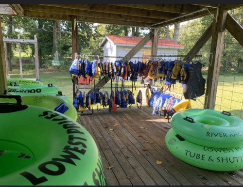A wooden shelter with colorful life jackets hanging and green inflatable tubes on the floor, surrounded by trees.