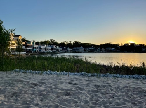 Sunset over a calm lake, with sandy beach in the foreground and charming houses lining the shore.