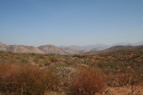 A panoramic view of rolling hills and mountains under a clear blue sky, with sparse vegetation in the foreground.