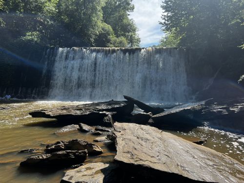 A serene waterfall cascades over rocks, surrounded by lush greenery and a bright blue sky.