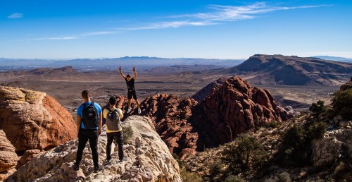 Three hikers stand on a rocky outcrop, with one celebrating atop a peak, overlooking a vast desert landscape.