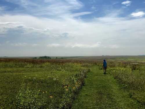 A person walks along a grassy path in a wide, open landscape under a blue sky with scattered clouds.