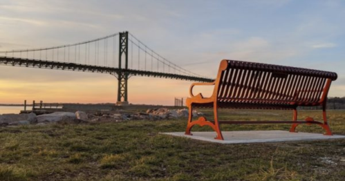 A wooden bench overlooks a river with a bridge in the background during sunset.