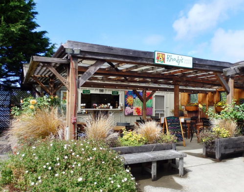 A cozy café with outdoor seating, surrounded by colorful flowers and greenery under a blue sky.