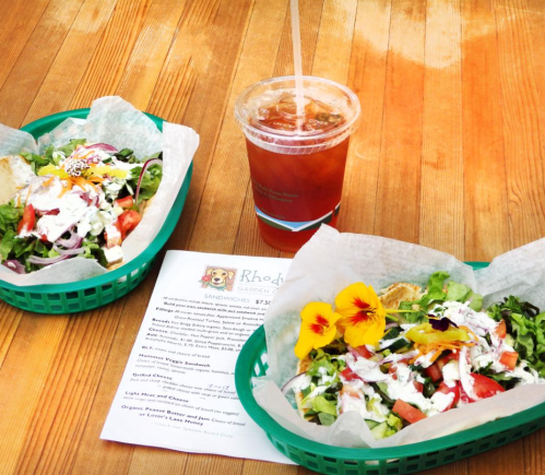 Two colorful salads in green baskets, a drink, and a menu on a wooden table, garnished with edible flowers.