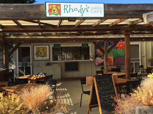 A cozy café entrance with outdoor seating, featuring a sign for "Rhody's Garden Café" and colorful flower decorations.