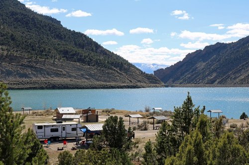 A scenic view of a lake surrounded by mountains, with an RV and picnic area in the foreground under a clear blue sky.