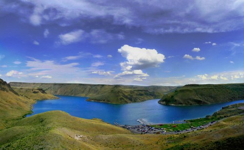 A panoramic view of a serene lake surrounded by green hills and a blue sky with fluffy clouds.