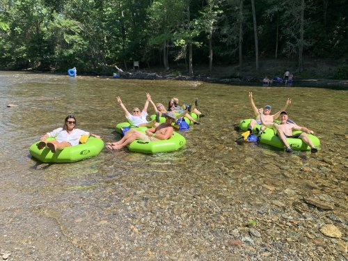 A group of six people relax on green inner tubes in a river, smiling and waving in a sunny, wooded setting.