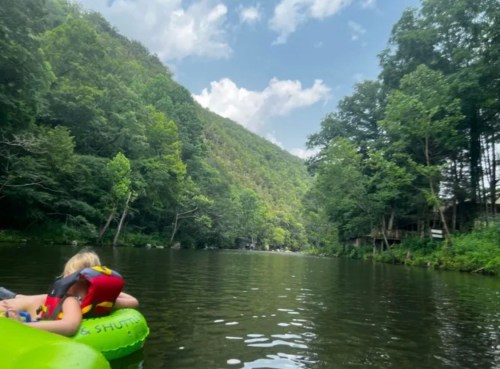 A person relaxes on a green float in a serene river surrounded by lush green mountains and trees under a blue sky.