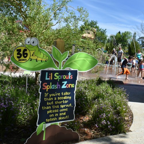 A sign for the "Lil Sprouts Splash Zone" with a height requirement of 36 inches, surrounded by a splash pad and children playing.