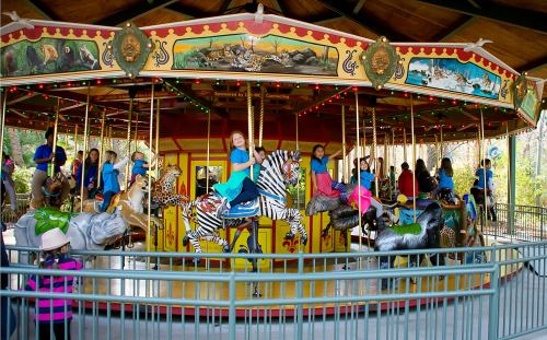 Children ride a colorful carousel featuring various animal figures, surrounded by festive decorations and greenery.