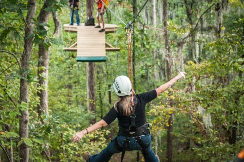 A person zip-lining through a forest, with a platform and two people visible in the background.