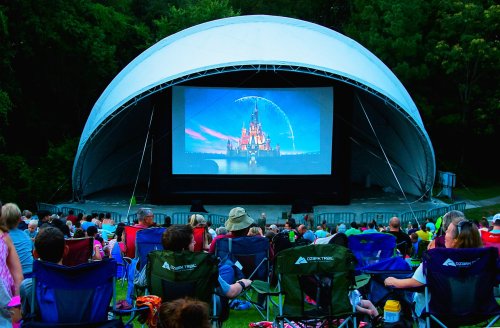 A crowd sits in folding chairs watching a movie on a large screen under a canopy in a park setting.