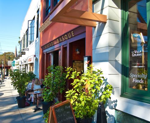 A vibrant street scene featuring a restaurant called "Bistro Basque" with outdoor seating and greenery.