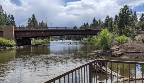A scenic view of a river with a bridge, surrounded by trees and greenery under a partly cloudy sky.