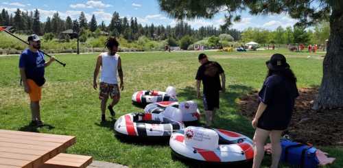 A group of people prepares inflatable rafts on a grassy area, with trees and a blue sky in the background.