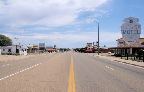 A wide, empty road stretches through a small town, lined with motels and businesses under a clear blue sky.