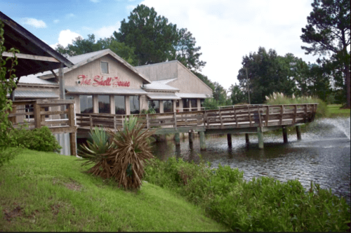 A rustic building with a wooden deck overlooking a pond, surrounded by greenery and trees under a cloudy sky.