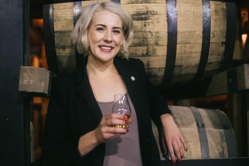 A smiling woman with short blonde hair holds a glass, standing beside wooden barrels in a distillery setting.