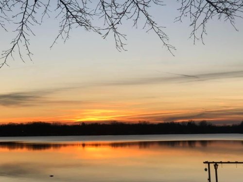 A serene sunset over a calm lake, with silhouettes of trees and vibrant orange and yellow hues in the sky.