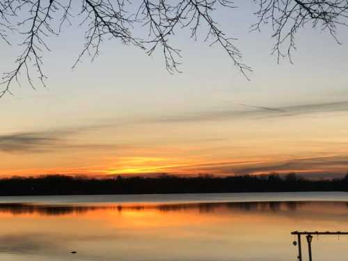 A serene sunset over a calm lake, with silhouettes of trees and vibrant orange and yellow hues in the sky.