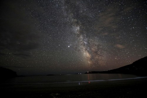 A stunning night sky filled with stars and the Milky Way over a calm beach, reflecting on the water.