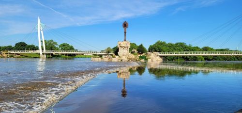 A scenic view of a river with a bridge, lush greenery, and a clear blue sky reflecting on the water's surface.