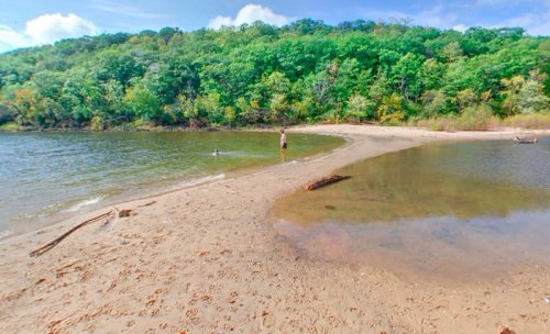 A sandy beach curves along a calm river, surrounded by lush green trees under a clear blue sky.