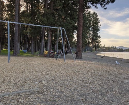 A sandy beach with a swing set, picnic tables, and trees, overlooking a calm lake under a cloudy sky.