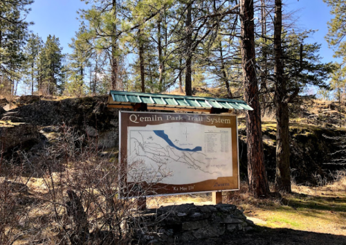 Trail map sign for Q'emlin Park Trail System, surrounded by trees and rocky terrain.