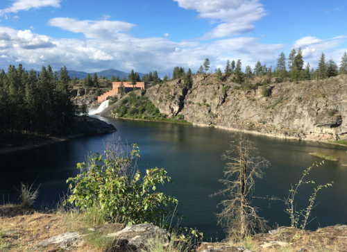A serene landscape featuring a calm river, rocky cliffs, and a waterfall, surrounded by trees under a blue sky.