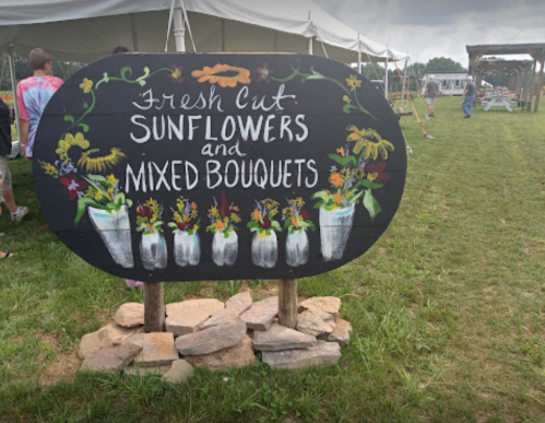 A sign reading "Fresh Cut Sunflowers and Mixed Bouquets" stands in a grassy area with a tent in the background.