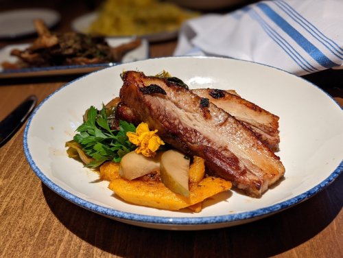 A plate of grilled pork belly served with garnished vegetables and a flower, on a wooden table.