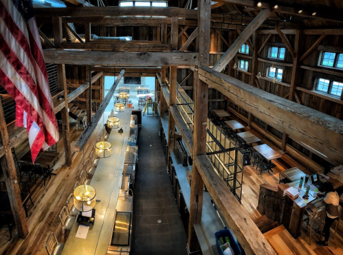 A spacious wooden interior of a barn-style restaurant with a long bar, tables, and an American flag hanging.