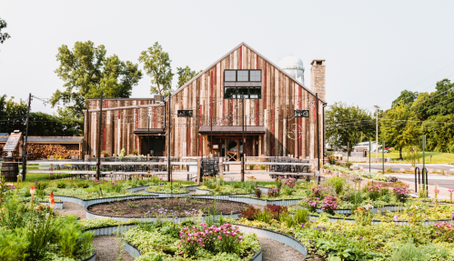A rustic barn-style building surrounded by lush gardens and flowers, with trees and a clear sky in the background.