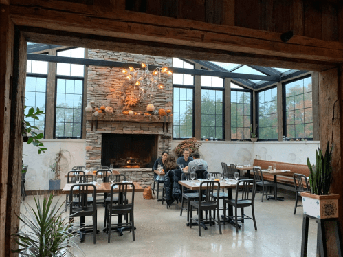 Cozy restaurant interior with a stone fireplace, large windows, and wooden decor, featuring tables and a couple dining.
