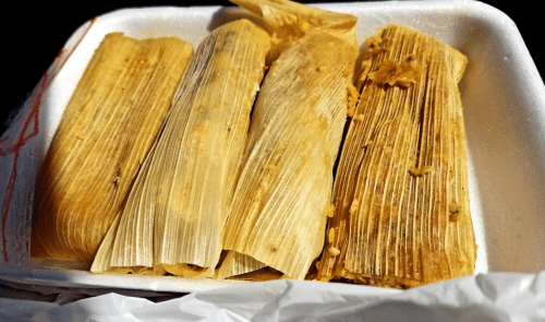 A takeout container with several tamales wrapped in corn husks, showcasing their golden-brown color.