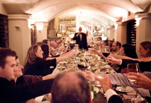 A group of people toasting with drinks at a long dinner table in a warmly lit restaurant.