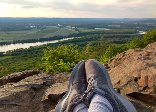 A pair of gray sneakers resting on a rocky ledge, overlooking a lush green valley and winding river below.