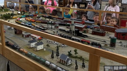 A model train display with children and adults observing, featuring various trains and a miniature landscape.