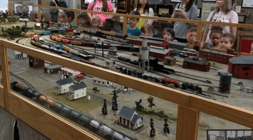 A model train display with children and adults observing, featuring various trains and a miniature landscape.